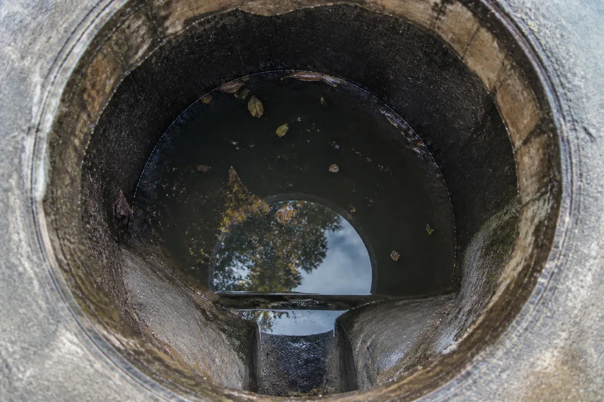 Regard d&rsquo;assainissement ouvert dans une cour caillouteuse, eau stagnante au-dessus de la cunette